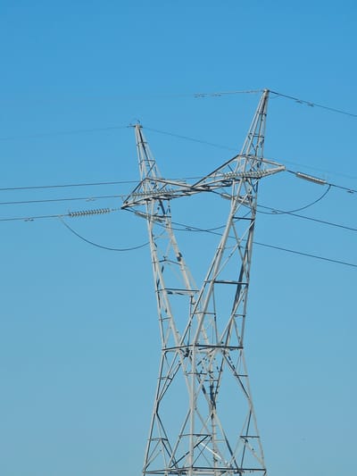 A solitary, weathered steel electricity pylon stands against a clear, bright blue sky. The pylon's lattice structure is visible in detail, with its crossbars and supporting beams forming a complex geometric pattern. Several thick power lines extend horizontally from the top of the pylon, disappearing into the distance. These lines are supported by insulators, which are also clearly depicted. The pylon itself is gray and appears to be of a standard design, likely used for transmitting high-voltage electricity. The scene is devoid of any surrounding landscape or other man-made structures, focusing solely on the pylon and the sky. The overall impression is one of industrial infrastructure set against a vast, unblemished sky. The location is stated as Cuarte de Huerva, Spain, though this context is not visually apparent from the image itself.