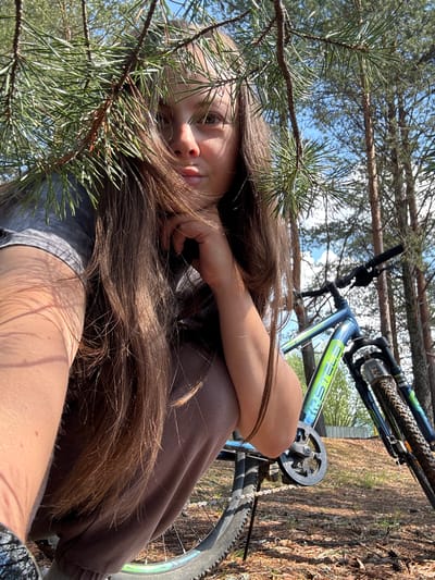 The image shows a young woman with long, brown hair reclining next to a bicycle in a wooded area. The woman is wearing a brown sweatshirt and has her head resting on her hand. A branch with pine needles is partially obscuring her face. The bicycle is positioned to the right of the woman. The location appears to be outdoors, with trees and a clear sky visible in the background. There is no information given in the picture that indicates that it was taken in Малая Рукавицкая, Russia.