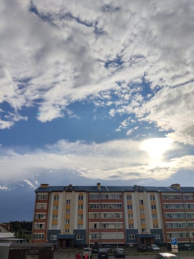 The image depicts a multi-story apartment building with a blue roof, located in Андрэеўшчынскі сельскі Савет, Belarus. The building features a facade with alternating sections of red brick and yellow and grey panels. Windows are visible on each floor, many with open or closed blinds. The sky above is partially cloudy with patches of blue sky and the sun is visible, creating a bright glare. In front of the building, there is a paved area with several parked cars, including a dark SUV, a sedan, and other vehicles. A blue parking sign with an arrow and "7M" is visible on the right side. A few individuals are standing in front of the building, some wearing red clothing. To the left of the apartment building, there are other structures and a line of trees in the distance, indicating a semi-urban or rural setting. The lighting suggests it is daytime, possibly late afternoon.
