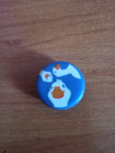 A blue round pin with cartoon-style white and orange geese is placed on a wooden surface. The pin is approximately in the center of the frame. The wooden surface has a visible grain pattern. There are no people or any other objects visible in the image. The location in the city of Promyshlennaya, Russia cannot be determined from the image.