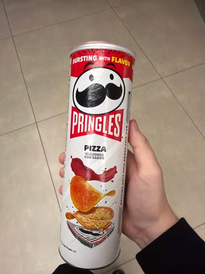 The image shows a hand holding a can of Pringles. The can is mostly white with red accents and features the Pringles mascot. The text on the can reads "BURSTING WITH FLAVOR" and "PRINGLES PIZZA FLAVORED CON SABOR". There is also text that reads "NOTHING HALF FULL ABOUT THIS CAN". The background appears to be a tiled floor. The lighting suggests an indoor setting, possibly a store or a home. The image is taken from a slightly elevated angle, looking down at the can and hand. There is no indication of the time of day, weather, or specific location beyond what is suggested by the Pringles product itself. The focus is on the Pringles can and the hand holding it.