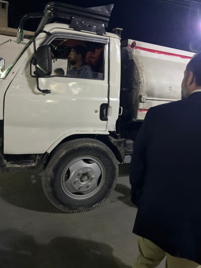 The image depicts a nighttime scene in Al-Kiswa, Syria, featuring a white utility truck and two male individuals. The truck, with a white cab and a white rear section (possibly a dump bed or tank) marked with a horizontal red stripe, is stationary on a dark asphalt or concrete surface. A male driver, with dark hair and a beard, is visible through the open driver's side window, wearing a dark shirt and looking forward. An orange object is partially visible within the cab behind him. A black roof rack is mounted on the cab. To the right of the truck, a second male individual is observed from behind, facing the truck. This individual wears a dark suit jacket and light-colored trousers. His right arm is slightly bent. The scene is illuminated by artificial light, with a bright light source or flare visible in the upper right quadrant of the image.