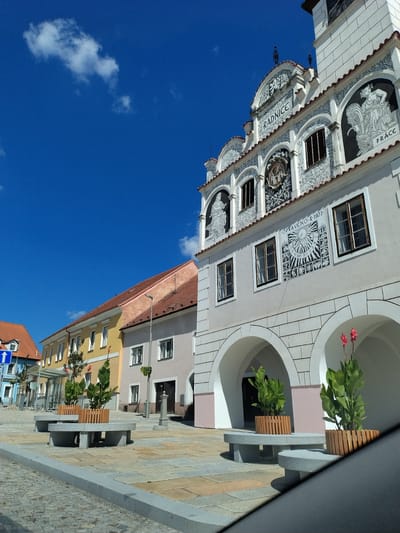 The image shows a town square with a white building that has ornate decorations and arched entrances in Volyně, Czechia. The building features detailed murals and text, including the word "RADNICE," which translates to "Town Hall," and "PRÁCE," meaning "Work." To the left of the town hall are several colorful, multi-story buildings with red tiled roofs. The ground is paved with cobblestones and flagstones. In the foreground, two stone benches with planters containing green plants and red flowers are placed in front of the town hall's arches. A clear blue sky with a few scattered clouds is visible above. The scene appears to be a sunny day in a historical European town.