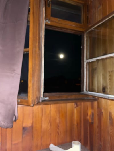An interior view looking out an open window at night shows a moonlit sky. The window frame is made of wood, as is the surrounding wall. A dark curtain hangs on the left side of the window. Outside, a blurred landscape is visible with a bright, full moon in the center of the frame. The scene suggests a room with wooden paneling and a table in the foreground with what appears to be a roll of toilet paper. The image is grainy and out of focus. The location in Jihlava, Czechia is not explicitly indicated by any visual cues in the image.