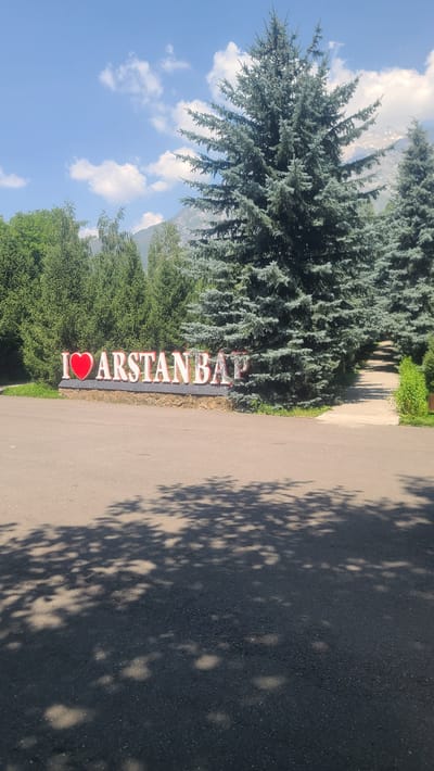 A large, three-dimensional sign reading "I ❤️ ARSTANBAP" is centrally positioned in front of a stone retaining wall. The sign's letters are white with red trim, and the heart symbol is red. Behind the sign, a dense grove of tall, dark green evergreen trees forms a backdrop. To the right of the sign, a paved path winds uphill and disappears among more trees. The foreground consists of a dark asphalt surface with dappled shadows from overhead foliage. The sky above is a clear blue with scattered white clouds, and in the far distance, the rugged peaks of mountains are visible, partially obscured by trees. This is a sign indicating the entrance or presence of Arstanbap, a village in Kyrgyzstan known for its walnut forests and scenic beauty.