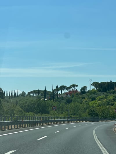 The image shows a highway in a rural, hilly area. The road curves to the right, with a metal guardrail on the left. The surrounding landscape is lush green with trees and vegetation. In the distance, there are some houses nestled on the hills, and a power line pylon. The sky is clear and blue. A speed limit sign of 100 is visible on the right side of the road. Based on the landscape and architecture, this scene is likely located in or near Impruneta, Italy.