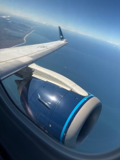 The image displays an aerial view from an aircraft window, showcasing the airplane's right wing, engine, and the external environment, presumed to be near the city of Unknown, Brazil. The wing is light grey/white, featuring visible panel lines and rivets, with partially obscured identification numbers or letters on its upper surface. A dark blue winglet, adorned with a colorful, possibly tropical-themed emblem, extends upwards from the wingtip. The engine nacelle is predominantly dark blue with a light grey inlet cowl and a distinctive bright blue horizontal stripe. Sunlight reflects prominently off the dark blue surface of the engine, illuminating various access panels and a yellow triangular warning symbol. Through the window, a vast expanse of calm, deep blue ocean is visible, bordered by a light-colored sandy coastline that separates the sea from a series of inland lagoons or bodies of water and distant landmass. The sky above is clear light blue, with a faint line of small cumulus clouds visible on the distant horizon. No people are visible in the scene. The overall lighting suggests a clear, sunny daytime environment.