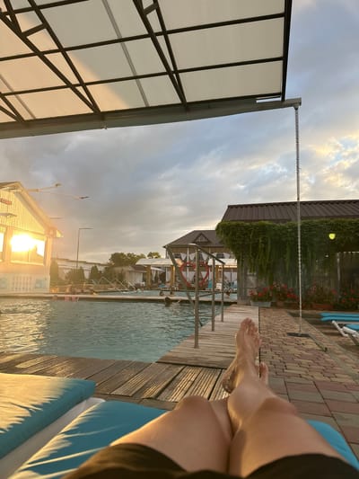 The image depicts a poolside scene with a person's legs and feet visible in the foreground, resting on a blue chaise lounge. The setting appears to be a resort or spa, likely in Yaroslavskaya, Russia, given the context. The sky is cloudy with hints of sunset colors. The pool itself is visible, with several people swimming and relaxing. A wooden deck surrounds the pool, and beyond that, there are buildings, one of which has ivy growing over its roof. The overall atmosphere is one of leisure and relaxation.