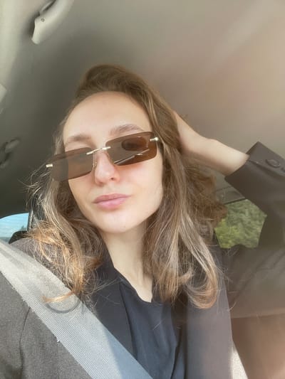 A young woman with long, light brown hair, wearing sunglasses and a black jacket, is photographed from the chest up in a car. She is touching her hair with her right hand. The image is taken from a low angle, looking up at her face. The background shows the interior of the car, including a seatbelt, and a blurred view of green foliage through the window. There is no indication of the city of Shud'ya, Russia, in the image.