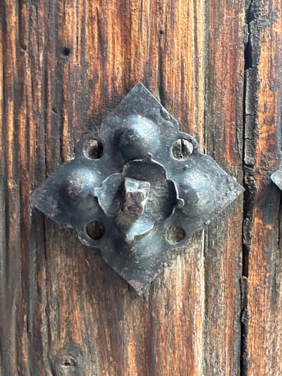 This is a close-up, eye-level, vertical photograph of an ornate, dark metal embellishment attached to rough, weathered, vertical wooden planks. The metal piece is diamond-shaped with decorative rounded points and features a central floral motif with a small, four-pronged metal clasp. It is affixed to the wooden surface with visible nail heads. The wood exhibits a textured grain with variations in color, ranging from light brown to dark brown and black, suggesting age and exposure. The overall scene appears to be a detail from an old door or gate in Huejotzingo, Mexico.
