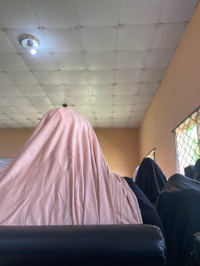 The image shows a person seated on a black chair, wearing black clothing and with a peach-colored blanket draped over their lap. The setting appears to be indoors, with white tiled walls and a tan ceiling. Visible through what seems to be a window with a wire mesh is some greenery, suggesting an exterior view. The location is given as Sabo, Nigeria.