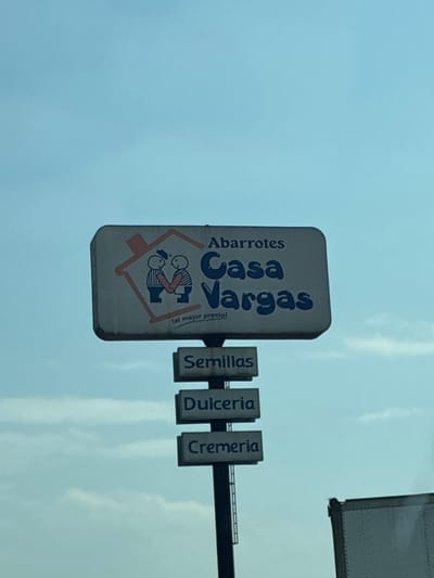 The image shows a sign for "Abarrotes Casa Vargas" in Tepeji de Ocampo, Mexico. The sign is white with blue lettering and features a logo depicting two figures inside a house shape. Below "Casa Vargas" it says "tal mejor precio!". Additional signs alongside list "Semillas," "Dulceria," and "Cremeria," indicating the store's offerings. The background is a light blue sky with some clouds. A portion of a truck or trailer is visible in the upper right corner. The sign is mounted on a pole or structure over what appears to be a roadway.