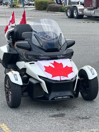 The image shows a three-wheeled vehicle, possibly a Can-Am Spyder, parked outdoors on asphalt. The vehicle is white and black, with a Canadian flag prominently displayed on its front. Two smaller Canadian flags are also attached to the vehicle's side. The vehicle appears to be on a paved surface, possibly a parking lot or street, with a yellow line visible in the lower right corner. In the background, a large red truck is partially visible, suggesting a commercial or industrial setting. A line of yellow and black bollards is also present, separating the area where the vehicle is parked from another space. There is some greenery in the background, indicating an outdoor setting. The lighting suggests it is daytime, and the surface of the vehicle and windshield shows some water droplets, possibly indicating recent rain or damp conditions. The overall scene suggests a display or gathering, given the prominent display of Canadian flags on the vehicle. No people are visible in the image. The word "LEV" is visible on a black banner in the background on the left side of the image.