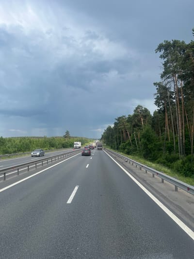 The image shows a view from a vehicle traveling on a multi-lane highway. The highway is paved and has clear white lane markings. A metal guardrail runs along the right side of the road, separating the highway from a dense forest of pine trees. The sky is overcast with dark, heavy clouds. Several vehicles are visible on the highway, including cars and trucks. In the distance, more trees and vegetation can be seen. The scene suggests a cloudy day on a highway bordered by a forest. There is no indication that this scene is located in Lukhovitsy, Russia.