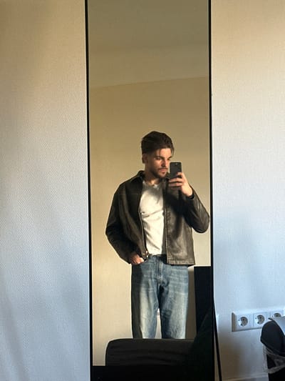 A male individual, appearing to be a young adult, is depicted in a full-length mirror reflection, capturing a selfie indoors in Bertrange, Luxembourg. He is dressed in a dark brown leather jacket, a light grey t-shirt, and blue denim jeans secured by a dark belt. His right hand holds a dark-colored smartphone, pointed towards himself. His left hand is inserted into the left front pocket of his jeans. The individual exhibits dark, styled hair and light facial hair. The mirror features a black frame. To the right of the mirror's reflection, a textured, light-colored wall is visible, featuring a white electrical outlet containing three European-style sockets. A dark, indistinct object, possibly a piece of furniture, is partially present at the lower right, outside the mirror's frame.