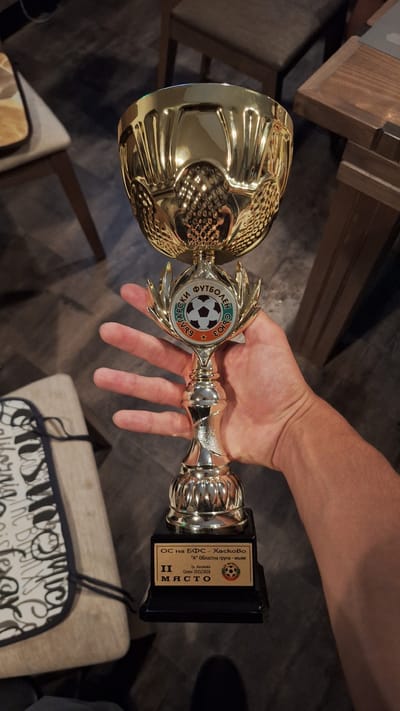 A hand is holding a gold and silver trophy. The trophy has a badge that says "ФУТБОЛЕН СЪЮЗ БЪЛГАРСКИ". The base of the trophy has a badge that says "ОС на БФС - XackoBo "А" Областна група - мъже Гр. Хасково Сезон 2025/2026 II МЯСТО". The location cannot be determined from the image.