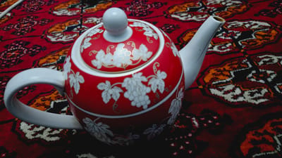 A red teapot with white grape and leaf designs sits on a red patterned rug. The teapot has a white handle and spout. The rug has a complex pattern in shades of red, orange, and white. The lighting suggests it is daytime.