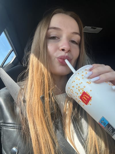 A young woman is in the passenger seat of a vehicle, seemingly drinking from a McDonald's cup with a straw. She has long blonde hair and is wearing a black leather jacket. The image shows details of the vehicle's interior, including the seatbelt. The location is unknown and can not be identified from the image.
