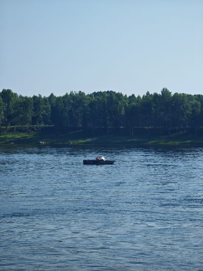 A lone small boat with two occupants is on a body of water. The boat is dark colored and appears to be an inflatable dinghy. One occupant is wearing a bright orange life vest. The boat has lettering on its side, which is mostly illegible, but appears to start with "SAL." A dense line of green trees forms the background, with a clear, light blue sky above. The water has small ripples on its surface. The scene is likely a calm day on a river or lake. The image has a high resolution and shows detail in the ripples of the water and the foliage of the trees. The overall impression is one of peaceful solitude.