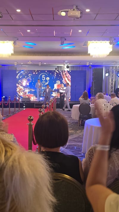 The image depicts an indoor event with a red carpet leading to a stage. A large screen displays a celebratory graphic with fireworks and the number "5" prominently featured, along with some text. On the stage, two men in suits are standing, appearing to be part of a presentation or award ceremony. To the right of the stage, a group of people are seated, some facing the stage and others turned away from the camera, suggesting an audience. The setting is illuminated by stage lighting and ambient room lights, creating a formal and festive atmosphere. The presence of a red carpet, stage, and screen suggests an organized event, possibly an anniversary celebration or a corporate gathering. The overall mood appears to be positive and celebratory. The location context provided is Parekklisia Community, Cyprus.