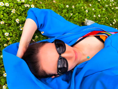 A person is lying on their back in a field of green grass and white daisies. They are wearing a bright blue hooded jacket, unzipped to reveal a colorful, patterned bra. The person is also wearing black sunglasses and a delicate gold necklace with a turquoise pendant shaped like a clover. One arm is bent at the elbow, with the hand resting on the back of their head. The image is taken from a high angle, looking down at the person. The environment is an outdoor grassy area, possibly a park or meadow, with a focus on the subject and their immediate surroundings. There is no indication of the city of Vecumnieki, Latvia.