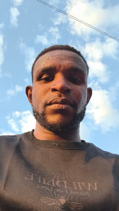A man with dark skin and a short beard gazes upward at the camera against a backdrop of a blue sky with scattered clouds. The lighting is intense, highlighting the features of his face. He wears a black t-shirt with a large, pale design covering much of the visible fabric. The perspective is a low angle, making the man appear to loom over the viewer. Two thin lines extend from the lower right, adding an element of visual texture.