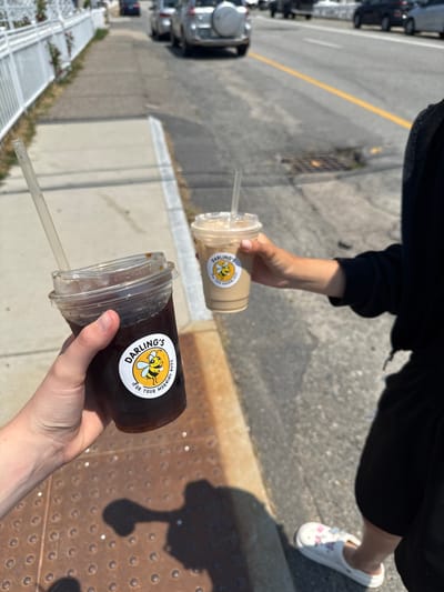 Two people are holding cups of coffee while walking on a sidewalk in Hampton, USA. The person on the left holds a dark-colored coffee in a plastic cup with a "Darling's" sticker featuring a bee. The person on the right holds a lighter-colored coffee, also in a plastic cup with a "Darling's" sticker. Cars are visible on the street in the background. The sidewalk is made of concrete and has a metal grate. A white fence runs along the left side of the sidewalk. The weather appears to be sunny.