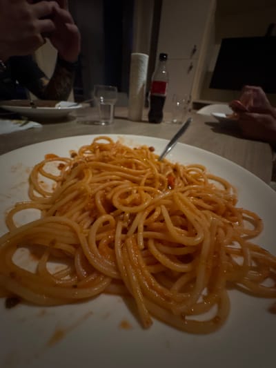 A plate of spaghetti with tomato sauce is on a table. There is a fork in the spaghetti. In the background, there is a bottle of Coca-Cola, a glass of water, and a roll of paper towels. There are also people in the background, but their faces are not visible. The scene appears to be a casual meal at home or in a restaurant. The image does not provide any information about the location in Deventer, Netherlands.