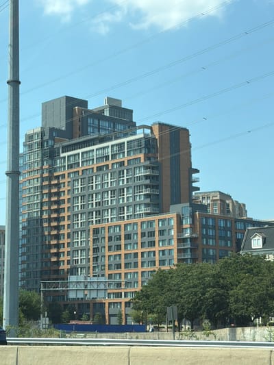 The image shows a tall, modern apartment building in Huntington, United States of America. The building has a complex geometric design with multiple sections of varying heights and colors, featuring a combination of glass windows, gray siding, and orange brick. Several balconies with glass railings are visible, extending from different levels. The building is situated in an urban environment, with a clear blue sky and some white clouds in the background. Several horizontal power lines stretch across the sky in front of the building. To the left, a large, gray utility pole is prominent. In the foreground, a concrete barrier and vegetation are visible, suggesting the vantage point is from a roadway or elevated structure. The overall scene depicts a contemporary urban landscape with a focus on the architectural details of the apartment building.