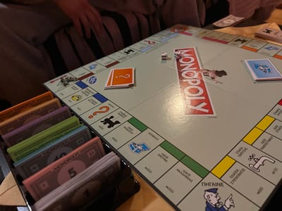 An overhead, close-up photograph depicts a Monopoly board game in progress or recently set up on a light-colored wooden table in Gerakas Municipal Unit, Greece. The game board is a Greek edition, featuring place names and instructions in the Greek language. Two white dice are visible on the board, displaying a "2" and a "3". A silver-colored key token rests near the upper left corner of the board. Stacks of orange "Chance" cards (with a question mark) and light blue "Community Chest" cards (with a character icon) are positioned on their respective spaces. A black plastic money tray contains organized stacks of Monopoly money in various denominations and colors, including 1, 5, 10, 20, 50, 100, and 500-unit bills. Partially visible in the upper left background are the legs and arm of an individual, dressed in striped pants and a light-colored garment, suggesting at least one player is seated at the table. A single 100-unit Monopoly bill lies separately on the table to the right. The scene is illuminated by warm, localized lighting.