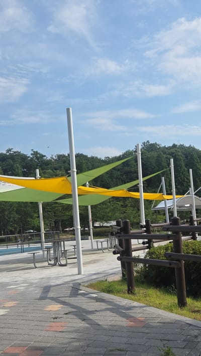 The image captures an outdoor seating area with a canopy of yellow and green fabric shades supported by white poles. Several metal picnic tables with attached benches are visible on a paved surface. In the foreground, a pathway made of interlocking gray, pink, and yellow rectangular bricks leads into the scene. A rustic wooden railing with multiple horizontal bars separates the paved area from a patch of green grass and dense foliage. The background is dominated by a thick, dark green forest under a light blue sky with scattered white clouds. The scene appears to be a public park or rest stop. The location is identified as Hwarang Recreation Area, Ansan City, South Korea. There are no people visible in the image.