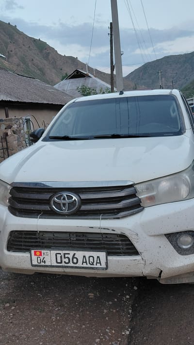 The image shows a front view of a white vehicle, likely a Toyota SUV, parked on a dirt road. The vehicle has a dented front bumper and grille. A license plate with the text "04 056 AQA" and "KG" with a red and yellow emblem is visible.

In the background, a rugged, mountainous landscape with sparse vegetation is visible under an overcast sky, suggesting a cloudy or potentially rainy day. A power line pole with wires runs diagonally across the scene. A portion of a building with a corrugated roof is also present to the left of the vehicle. The scene appears to be in a rural or mountainous region, consistent with the provided location context of Ak-Terek, Kyrgyzstan. There are no people visible in the image. The overall impression is that of a vehicle in a somewhat remote and possibly rugged environment.