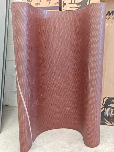 The image shows a large, curved piece of what appears to be a reddish-brown textured material with a subtle pattern of small dots. The material is bent into a U-shape, with its edges facing the viewer and its central section curving away. The surface has a slightly glossy finish and some visible scuff marks and streaks, indicating it might be a protective film or a material used in manufacturing or construction.
Behind the curved object, there are several cardboard boxes. One box on the right has a large black graphic, possibly a logo or illustration, and some faint text that is difficult to read clearly, but the letters "M" and "P 80" are partially visible. On the top of some of the boxes, there is printed text that reads "SAIT" in capital letters, repeated several times.
The setting appears to be an indoor industrial or workshop environment. The floor is concrete, and a white brick wall is visible on the far left, partially obscured by a vertical element that could be a doorway or a large piece of equipment. The lighting suggests it is daytime, but no specific time of day can be determined. The overall impression is of a material or product stored or awaiting use in a commercial or industrial space in Settimo Torinese, Italy. There are no people visible in the image.