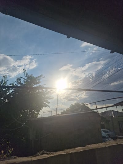 This is a photograph taken from a low angle, looking upwards towards the sky. The image is oriented such that the sky occupies the majority of the frame, with structures and foliage framing the scene. The sun is visible, bright and slightly obscured by clouds, indicating it is daytime. The sky is a vibrant blue, dotted with fluffy white clouds. Several power lines crisscross the sky, supported by poles which are visible on the right side of the frame. To the left and right edges of the image, dark, silhouetted structures create a sense of enclosure. On the right, the top of a building or wall is visible, with some roofing material and what appears to be a tarpaulin. In the lower right portion of the frame, the dense foliage of a tree is present, its leaves illuminated by the sun. The overall impression is of a sunny day, possibly late morning or early afternoon, viewed from a street or courtyard in a populated area, suggested by the presence of overhead wires and buildings. The lighting suggests a bright, clear day, though the contrast between the bright sunlit sky and the shadowed structures indicates varied lighting conditions. There is no visible text in the image. The scene appears peaceful and everyday.