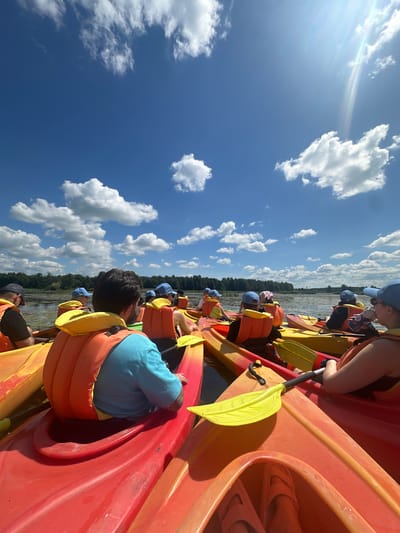 A group of people are in kayaks on a body of water, likely a lake or river, under a blue sky with scattered white clouds. The kayaks are colorful, primarily red and yellow. Many of the individuals are wearing orange life vests and blue caps. The foreground shows the close-up view of several kayaks, with paddles visible. In the middle ground and background, more kayaks are present, and a line of trees forms the shoreline. The sun is shining brightly, casting some glare on the water and sky, suggesting it's a sunny day. The scene appears to be a recreational activity, possibly a guided tour or group outing. The location is identified as the Валожынскі сельскі Савет, Belarus.