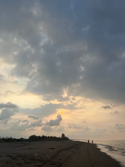 The image depicts a tranquil coastal scene at what appears to be sunset or late afternoon, characterized by soft, golden light. The location is Angkleng Barat, Indonesia.
The sky occupies the majority of the frame, filled with a mix of dense, dark gray clouds on the left, gradually transitioning to lighter, warm-toned clouds on the right, illuminated by the setting sun. The sun itself is visible on the right side of the sky, partially obscured by clouds, casting a bright, circular glow and streaking rays through the cloud cover. A very small, dark object, possibly a bird, is faintly visible in the upper left portion of the sky.
The lower and right-hand side of the image features a sandy beach meeting the ocean. Waves are visible breaking gently onto the shore, creating a foamy white line. The sand is a dark, wet color, with visible footprints and tire tracks suggesting recent activity. Along the far edge of the beach, a dense line of trees forms the horizon, with hints of distant structures or land beyond.
Two silhouetted figures are seen walking along the beach near the waterline, heading into the distance. One individual is wearing red clothing, while the other is in lighter attire. Their small scale against the expansive sky and beach emphasizes the vastness of the environment.
The overall mood is serene and peaceful, capturing a quiet moment at the end of the day by the sea. There is no visible text in the image.