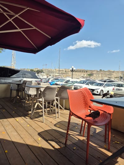 This image depicts an outdoor seating area at a waterfront cafe in Ta' Xbiex, Malta. The foreground features a wooden deck with scattered fallen yellow leaves. Several tables with chairs are arranged on the deck, with a large red umbrella providing shade. Two bright orange plastic chairs are prominently placed in the right foreground, with one occupied by a bird. In the mid-ground, more tables and chairs are visible, alongside a grey SUV and other parked cars. Beyond the seating area, a marina is visible with numerous boats and yachts docked. In the background, a historic walled city, likely Valletta, is situated on a hill overlooking the harbor, with tall mast-like structures of yachts and cranes also present. The sky is clear blue with a few scattered white clouds. The scene is illuminated by bright sunlight, casting distinct shadows from the furniture onto the deck.
