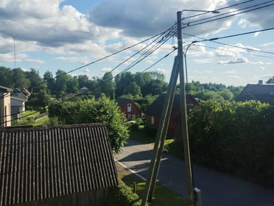 The image depicts a rural landscape, likely in Kanepi alevik, Estonia, during what appears to be a sunny afternoon with scattered clouds. The foreground is dominated by a corrugated, dark-colored roof of a building. To the right, a wooden utility pole with numerous overhead wires runs vertically through the frame. Beyond the roof and the pole, a paved road curves gently into the distance, bordered by green grass and hedges. Several red-roofed houses are visible among dense green trees, contributing to the idyllic, residential setting. The light suggests it is daytime, possibly late afternoon due to the shadows cast by the trees and utility pole. No people are visible in the scene. The overall impression is one of a tranquil, everyday rural environment.