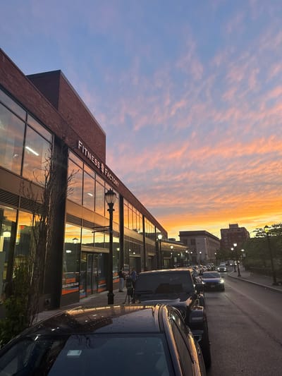 The image depicts a street scene in Yonkers, United States of America, at dusk. The sky is filled with colorful clouds ranging from orange to pink and blue. On the left side of the street, a brick building with large windows displays a sign that reads "FITNESS FACTORY." Reflections of the sunset are visible in the building's windows. A street lamp stands in front of the building. Several cars are parked along the street, and a few are in motion. The street is paved, and there are sidewalks with trees and other lamps. In the background, other buildings and trees can be seen.