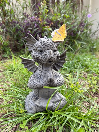 An outdoor, eye-level, medium shot captures a gray, stone garden figurine of a young dragon. The dragon is seated with its hands clasped in front of its chest and is facing forward. Its wings are folded against its back. A yellow butterfly with black markings is perched on the dragon's head, resting on a small white flower. The dragon figurine is surrounded by lush green grass and some foliage. In the background, blurred purple and green plants create a natural setting. The image appears to be taken in Riverview, United States of America, based on the provided location.