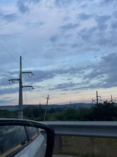 The image depicts a roadside view from a moving vehicle in Krymsk, Russia, with a focus on the sky and surrounding landscape. The sky is filled with a mix of gray and pale blue clouds, suggesting either early morning or late evening light. In the foreground, the side-view mirror of a white car is visible on the left, reflecting the cloudy sky and the car's interior. A metal guardrail runs horizontally across the middle of the frame, separating the road from the embankment. Beyond the guardrail, a lush green landscape with trees and rolling hills stretches into the distance. Several electrical transmission towers and power lines are scattered throughout the scene, indicating the presence of infrastructure. The overall impression is one of a rural or semi-rural setting under a dramatic sky.