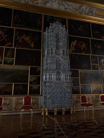 The image depicts an ornate interior scene, likely a historical palace or museum, featuring a tall, blue and white tiled stove as the central object. The stove is intricately decorated with patterns and architectural details, resembling columns and arched recesses. It stands against a wall covered with numerous framed paintings, arranged in a grid-like fashion. The paintings appear to be historical or religious in nature, with dark palettes and figurative subjects.

The floor is polished wood, reflecting the ambient light and exhibiting a geometric pattern. In the foreground, several opulent chairs with red velvet upholstery and gilded frames are visible, suggesting a reception or viewing area. These chairs are separated by golden stanchions and ropes, indicating a barrier for visitors. The walls also feature decorative gilded elements, particularly above the chairs. The lighting is subdued, creating a sense of grandeur and historical atmosphere.

Based on the architectural style, the tiled stove, and the overall opulent decoration, this scene is likely located within a significant historical building in Alexandrovskaya, Russia. Such interiors are characteristic of palaces like the Catherine Palace or similar imperial residences, which often house extensive art collections and elaborate decorative features.
