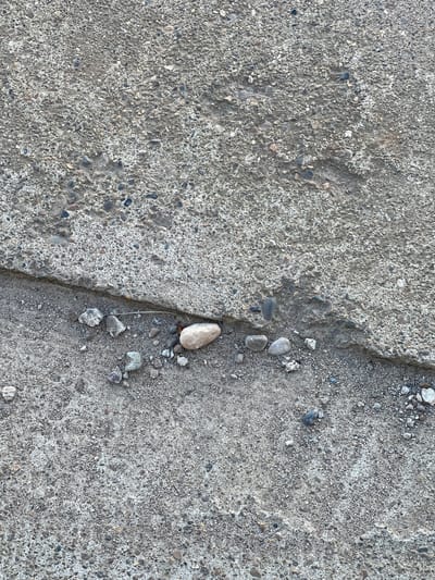 The image presents a close-up, overhead view of a cracked concrete surface in Бизяки, Russia. No people or discernible activity are present. The primary subjects are the concrete ground and various small stones and gravel embedded within it and along a prominent fissure.

A significant, irregular crack runs roughly down the center of the frame, dividing the concrete surface into two sections. This crack is wider and more jagged in the middle and lower portions, becoming slightly narrower towards the top. Within and alongside this crack, several loose stones and pebbles are visible. Notably, a light-colored, elongated stone rests within the crack near the center-right, surrounded by smaller, darker gravel.

The concrete exhibits a rough, aggregate texture, with numerous small, multi-colored stones (grays, browns, whites) exposed on its surface, indicating wear or a specific finish. The dominant color throughout the image is a range of grays, with subtle variations from the embedded aggregates and shadows within the crack. The lighting appears to be natural daylight, casting soft shadows that highlight the texture and depth of the crack. There is no visible text. The scene captures the aged and weathered detail of a paved surface.