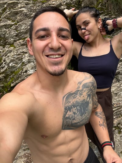 The image depicts a selfie of a shirtless man with a large chest tattoo, lying on a textured surface covered in moss and lichen, possibly a rock. He is smiling at the camera. Behind him, a woman wearing a dark tank top and brown pants is lying on her back, sticking her tongue out. She has a tattoo on her chest and what appears to be a strip of tape on her nose. The location appears to be outdoors, possibly in nature, based on the rock-like surface. There is no explicit information to place this photo specifically in Medven, Bulgaria.