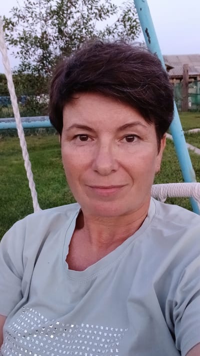 A woman with short, dark hair and fair skin is seated on a swing in an outdoor setting. She is wearing a light blue t-shirt adorned with silver rhinestones. The background features a green lawn, trees, and a rustic wooden structure. The lighting suggests it is either early morning or late evening. The woman is looking directly at the camera with a slight smile.