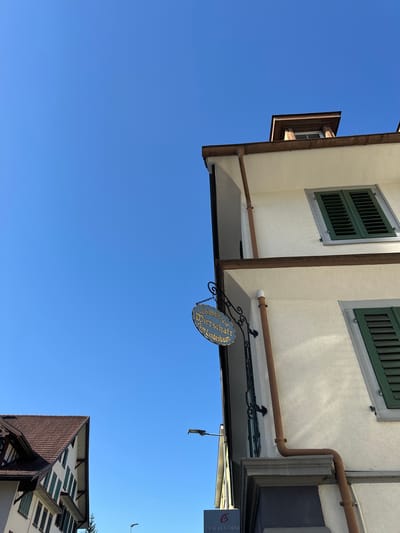 A low-angle, close-up shot captures the side of a white building with green shutters under a clear blue sky. A sign with gold lettering hangs from a decorative black metal bracket. The sign reads "Alkoholfreie Wirtschaft Zum Lindenbaum." To the left, a portion of another building with a brown tiled roof and dark trim is visible. The scene appears to be in Wald, Switzerland.