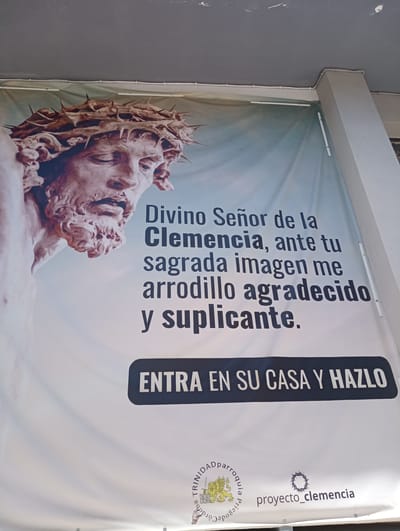 The image is a close-up shot of a large banner featuring a religious motif. The banner displays a partial depiction of Jesus Christ wearing a crown of thorns, rendered in a statue-like style with a pale, stony texture. To the right of the image of Jesus, there is text in Spanish. The top lines of text read "Divino Señor de la Clemencia, ante tu sagrada imagen me arrodillo agradecido y suplicante." Below this, a dark horizontal bar contains the text "ENTRA EN SU CASA Y HAZLO" in white capital letters. At the bottom of the banner, there are two logos and some smaller text. One logo features a lion and a shield with text, and the other is a crown of thorns. The text below these logos appears to be "proyecto_clemencia" and "parroquia_trinidad_priego_de_cordoba". The banner is mounted outdoors, possibly on the exterior of a building, with a grey ceiling or overhang visible at the top and a concrete pillar on the right side. The overall impression is that this is an advertisement or message from a religious institution, specifically the Parish of Trinidad in Priego de Córdoba, Spain.