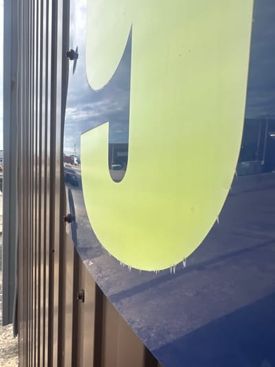 The image is a close-up, angled shot of a large, light green letter "P" set against a dark blue background. The letter appears to be made of a reflective material, as it mirrors the sky and surrounding environment, which seems to be outdoors. The "P" has a slightly distressed or worn edge along its curved portion, with small, jagged white protrusions. Below the letter, a metallic structure with horizontal ridged lines is visible, secured by several dark-colored rivets or fasteners. One fastener is prominently displayed on the left side of the frame, attached to the blue background and protruding slightly over the metallic structure. Another fastener is visible further to the right, closer to the bottom of the letter. The overall impression is that of an outdoor sign or logo, possibly on a vehicle or building, with a slightly weathered appearance. The lighting suggests it could be daytime, with bright, reflected light. There is no visible text in the image.