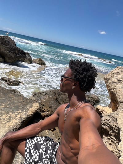 A man is sitting on rocks along the coast on a sunny day. The sea is rough and blue with white waves crashing against the rocks. The sky is clear and blue with a single small cloud. The man is wearing dark glasses and a silver necklace. He has black hair. He is wearing black and white checkered shorts. The location is Llucmajor, Spain.