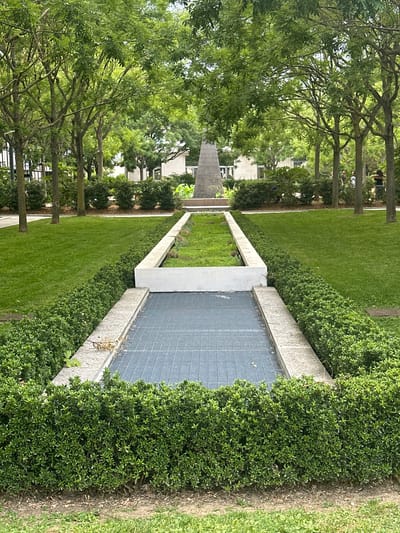 This is a photograph of a park in Enghien-les-Bains, France. The park is lined with trees and has a manicured lawn. In the center of the image, there is a pathway with a grated metal surface, bordered by hedges and concrete structures. At the far end of the pathway, a large, pointed monument stands. Several people are visible in the background, walking amongst the trees. The overall scene suggests a tranquil and well-maintained public space.